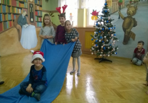 Świątecznie udekorowana sala. Troje dzieci tzryma niebieski kocyk na którym siedzi chłopiec w mikołajowej czapce.
